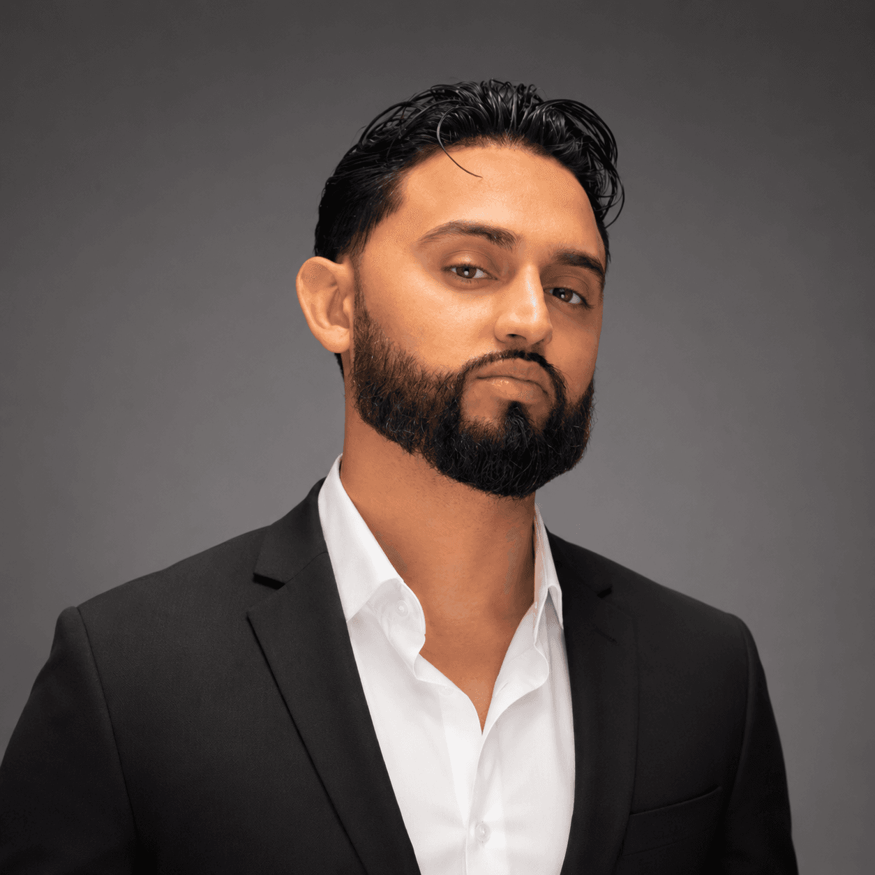 Bearded man in a black suit and white shirt against a solid dark gray background.
