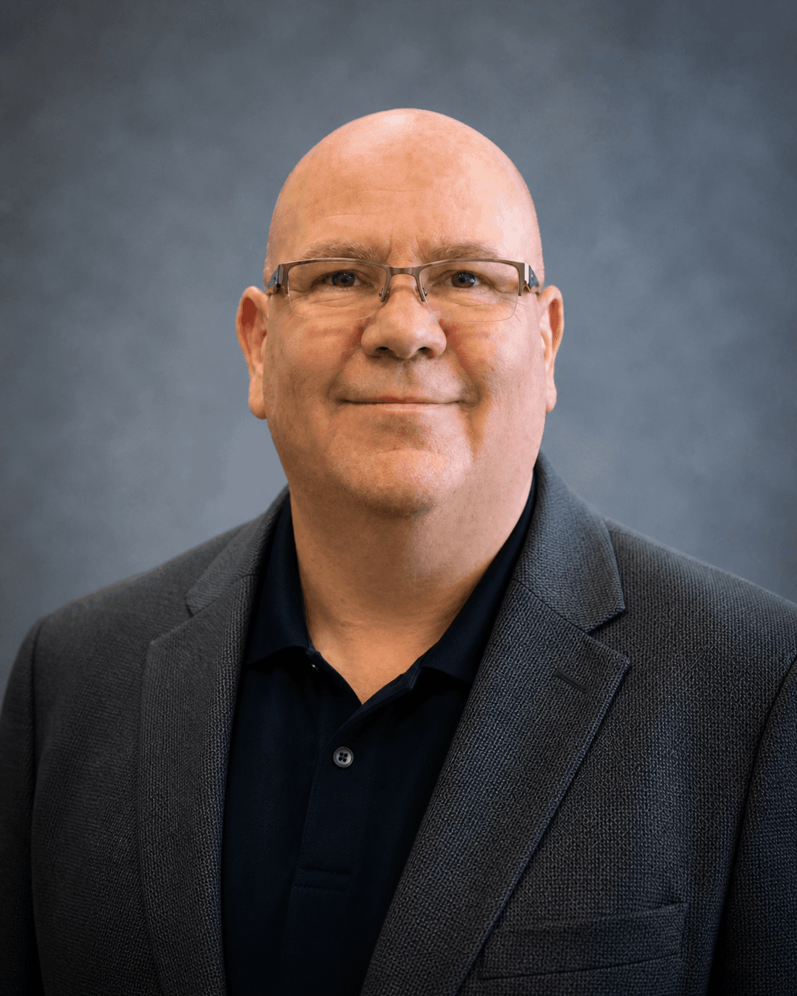 Smiling bald man with glasses wearing a blue polo shirt and grey textured blazer.
