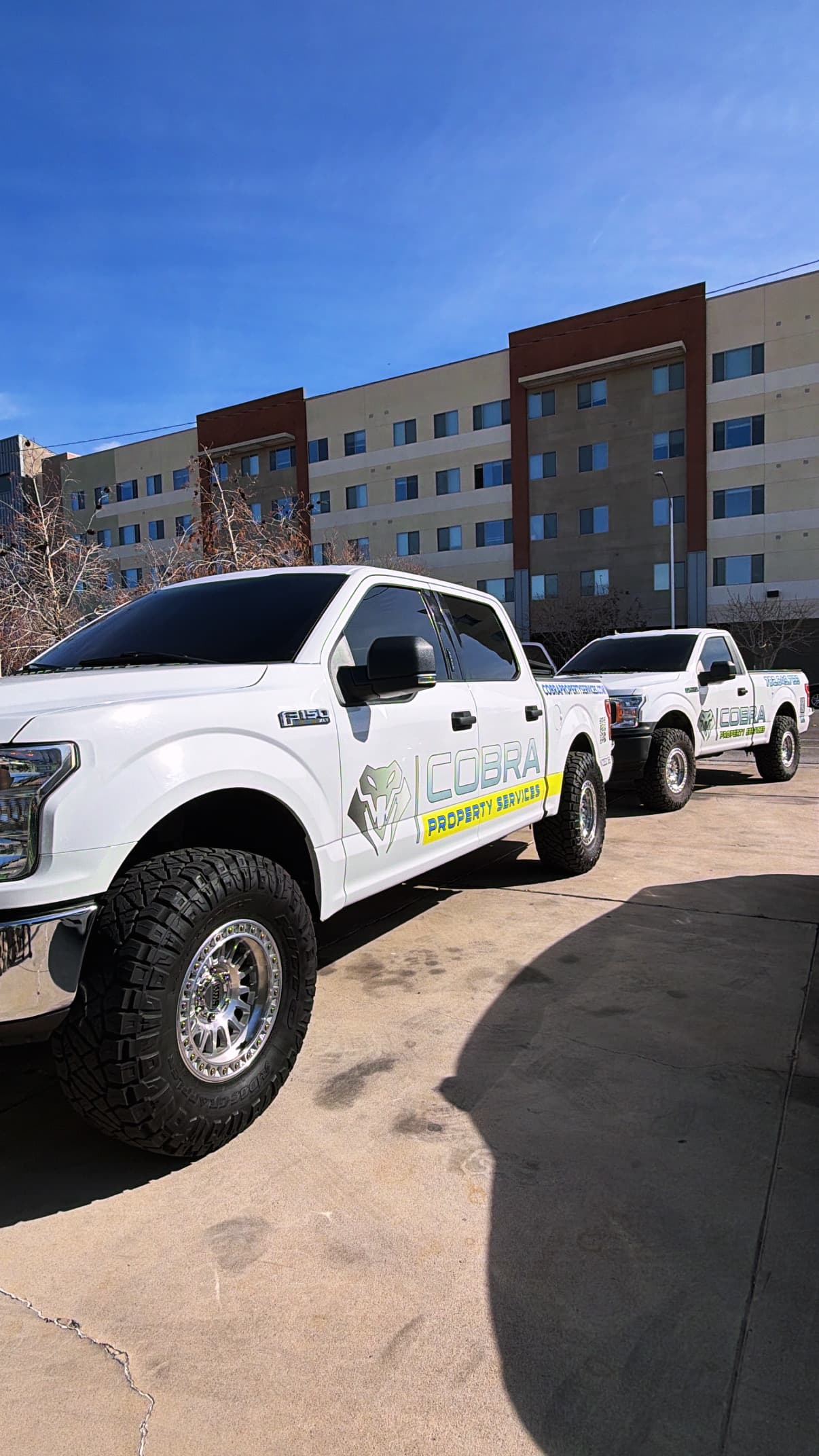 Two white Cobra Property Services trucks with off-road tires parked outside a multi-story building.
