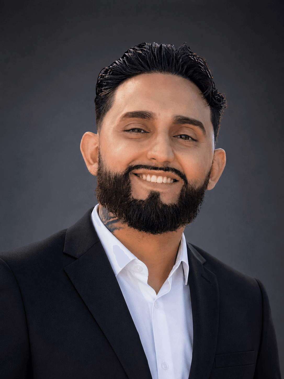 Smiling man with dark hair, a beard, and neck tattoo wearing a black suit jacket.