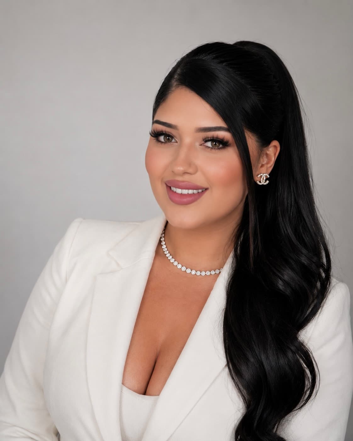 Smiling woman with long black hair, wearing a white blazer, diamond necklace, and earrings.